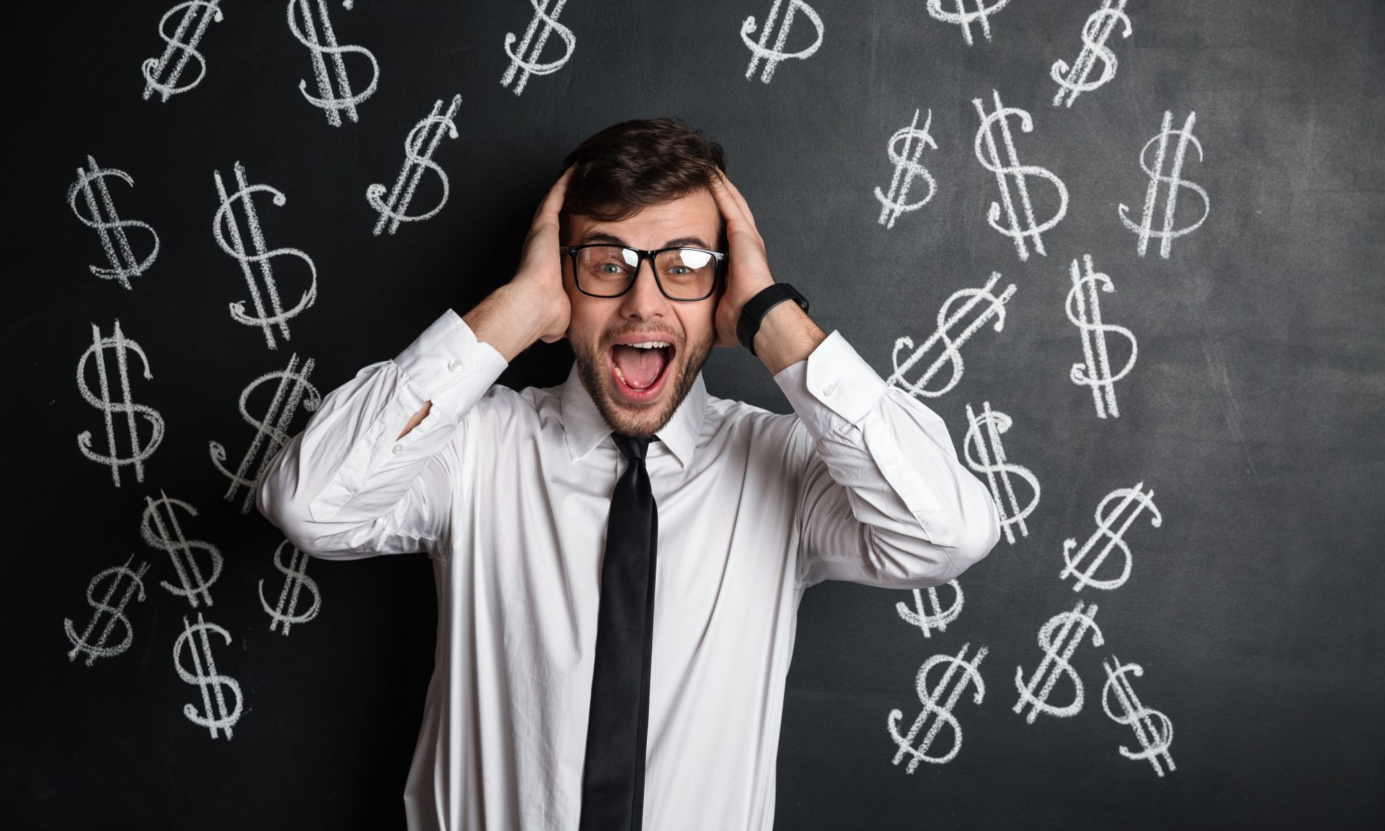 Overjoyed young bearded man in glasses holding his head, looking at camera, isolated over blackboard with dollars symbol, showing how confusing navigating a rent rise can be