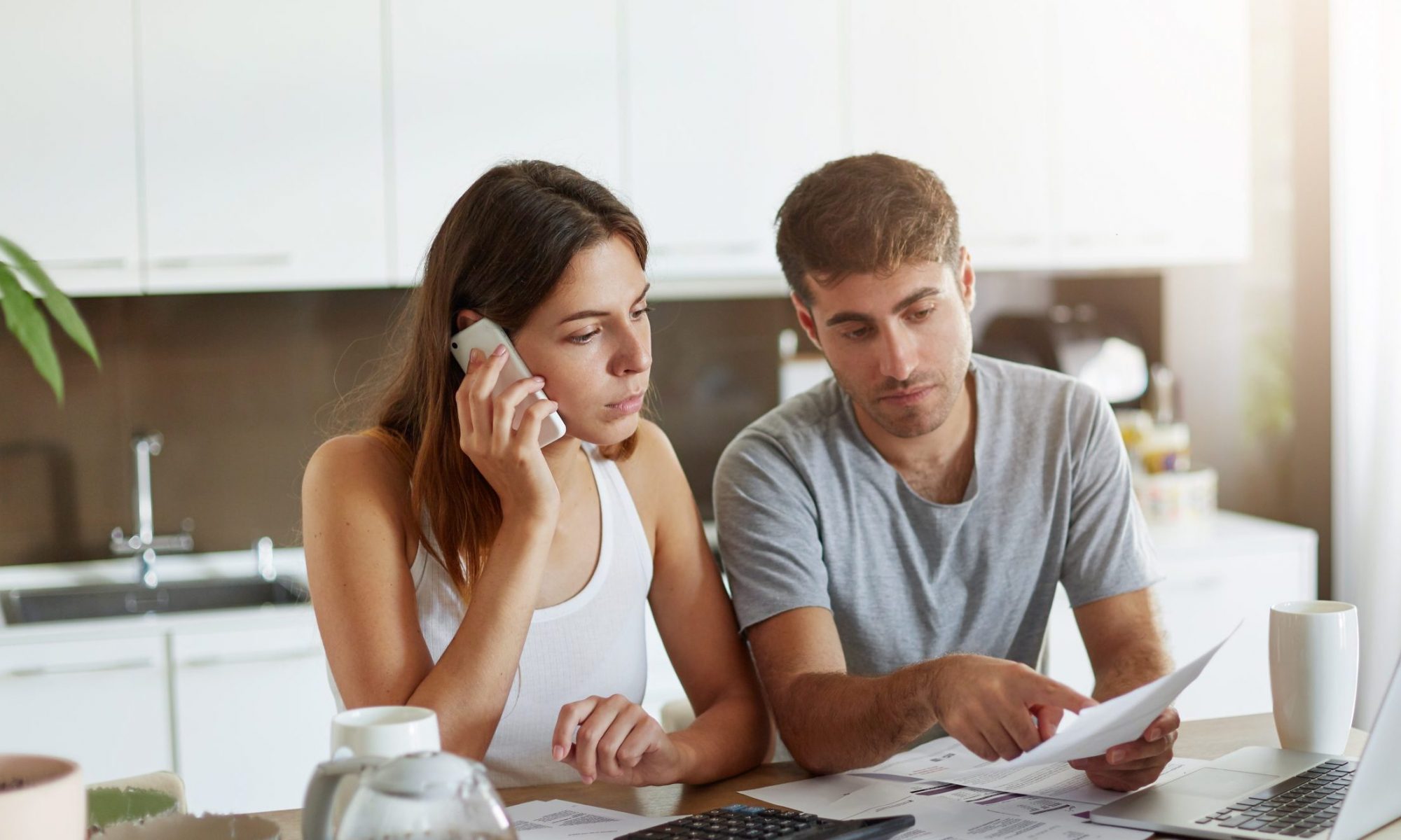 Young businessman pointing with finger at document, trying to expain something to his wife who is speaking over smart phone. Couple reviewing their bank accounts and calculating annual mortgage figures